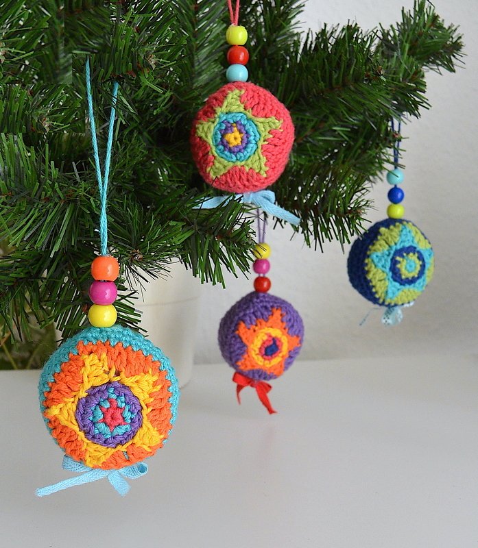 Vier farbenfrohe gehäkelte Weihnachtskugeln mit Perlen und Schleifen an einem Tannenbaum.