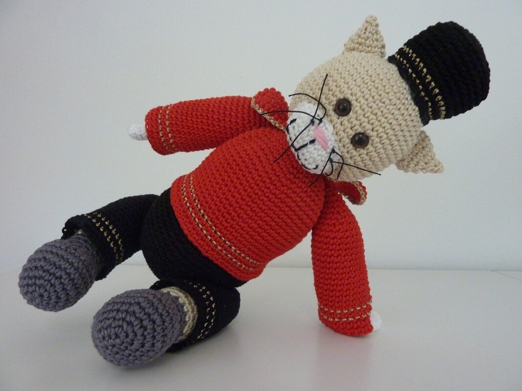 Beigefarbene gehäkelte Amigurumi-Katze mit roter Jacke, schwarzen Hosen und schwarzer Mütze