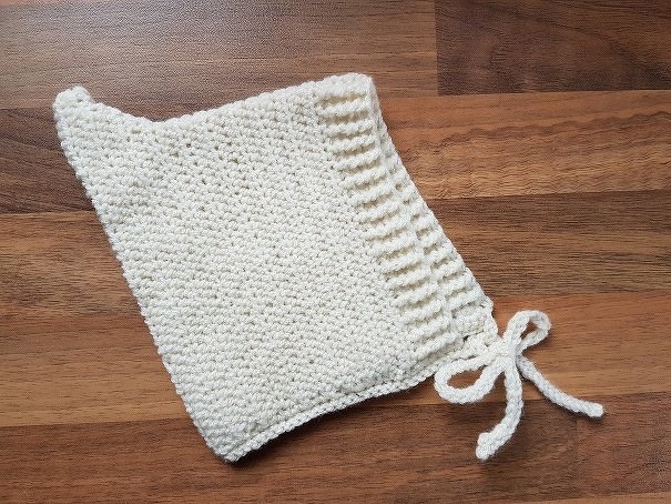 Cremefarbene gehäkelte Babymütze mit Bindebändchen auf Holztisch