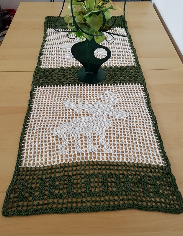 Gehäkelter grüner und weißer Tischläufer im Filetmuster mit Rentiermotiv auf einem Holztisch, dekoriert mit einer grünen Vase.
