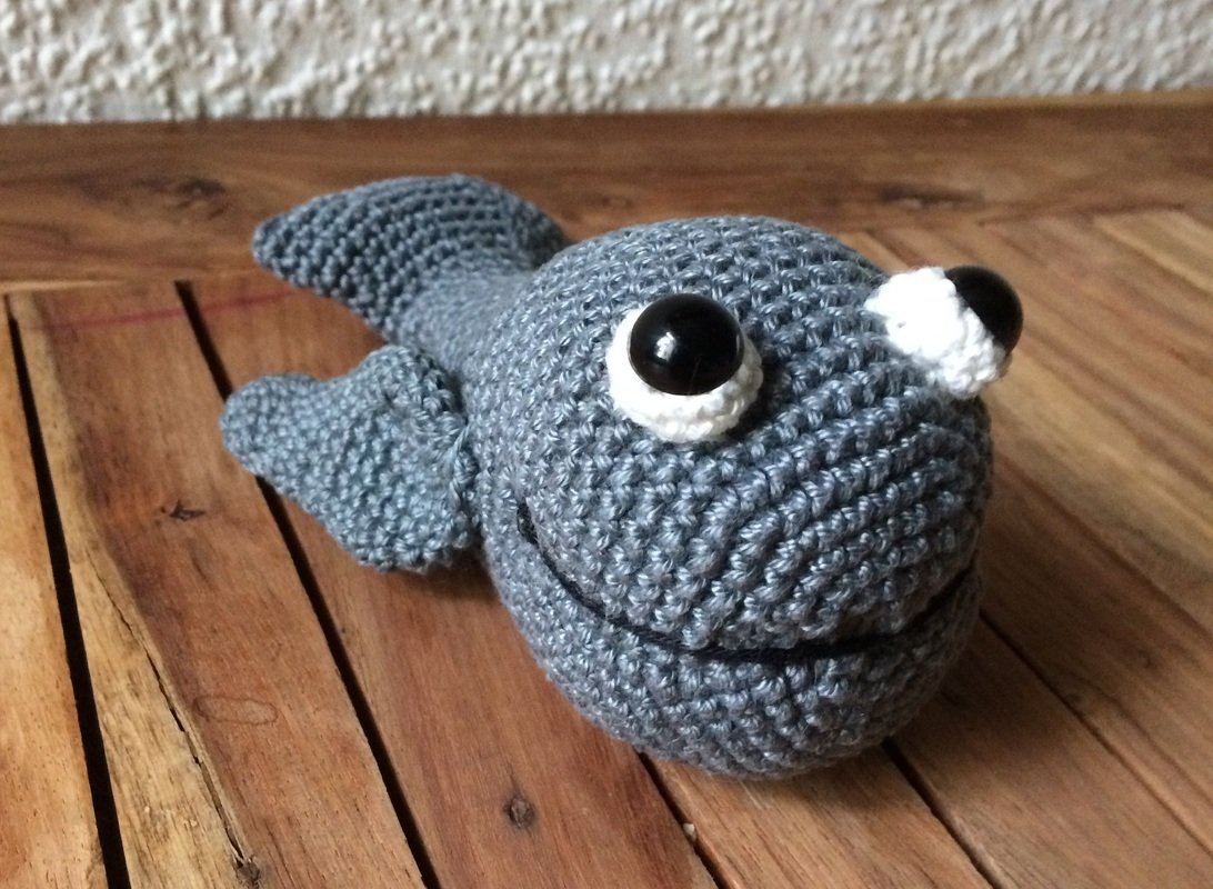 Gehäkelter grauer Amigurumi-Fisch mit schwarzen Sicherheitsaugen auf Holzuntergrund
