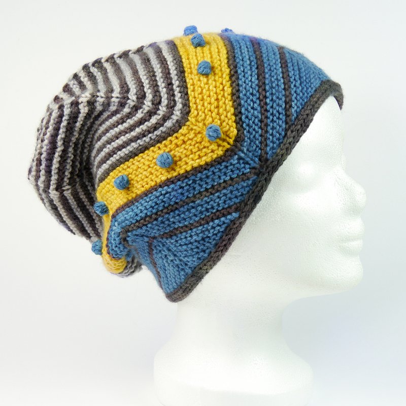 Locker fallende, gestrickte Beanie‑Mütze mit blauen, gelben und grauen Streifen und blauen Noppen auf weißem Schaufensterkopf.