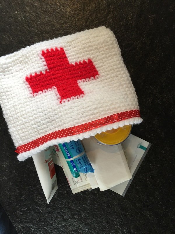 Weißes gehäkeltes Täschchen mit rotem Kreuz und gepunktetem roten Band, geöffnet mit sichtbaren Packungen und Papieren