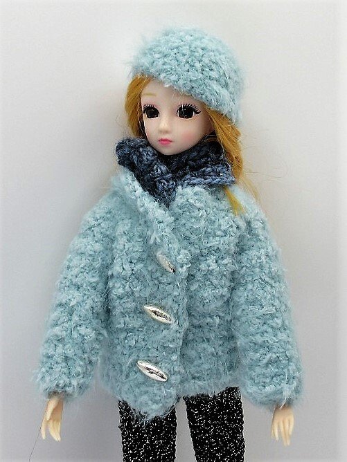 Puppe in gehäkeltem Winteroutfit: hellblaue Jacke, Mütze, dunkelblauer Schal und dunkle Hose.