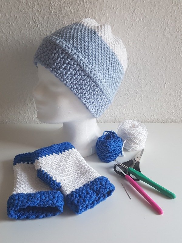 Gehäkelte Mütze und Stulpen in Blau-Weiß mit Garnknäueln, Häkelnadeln und Schere auf weißer Fläche