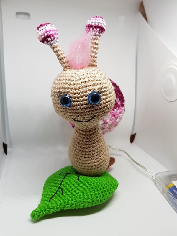 Gehäkelte Schnecke mit rosa Haarschopf, blauen Augen und rosa-violettem Gehäuse auf grünem Häkelblatt