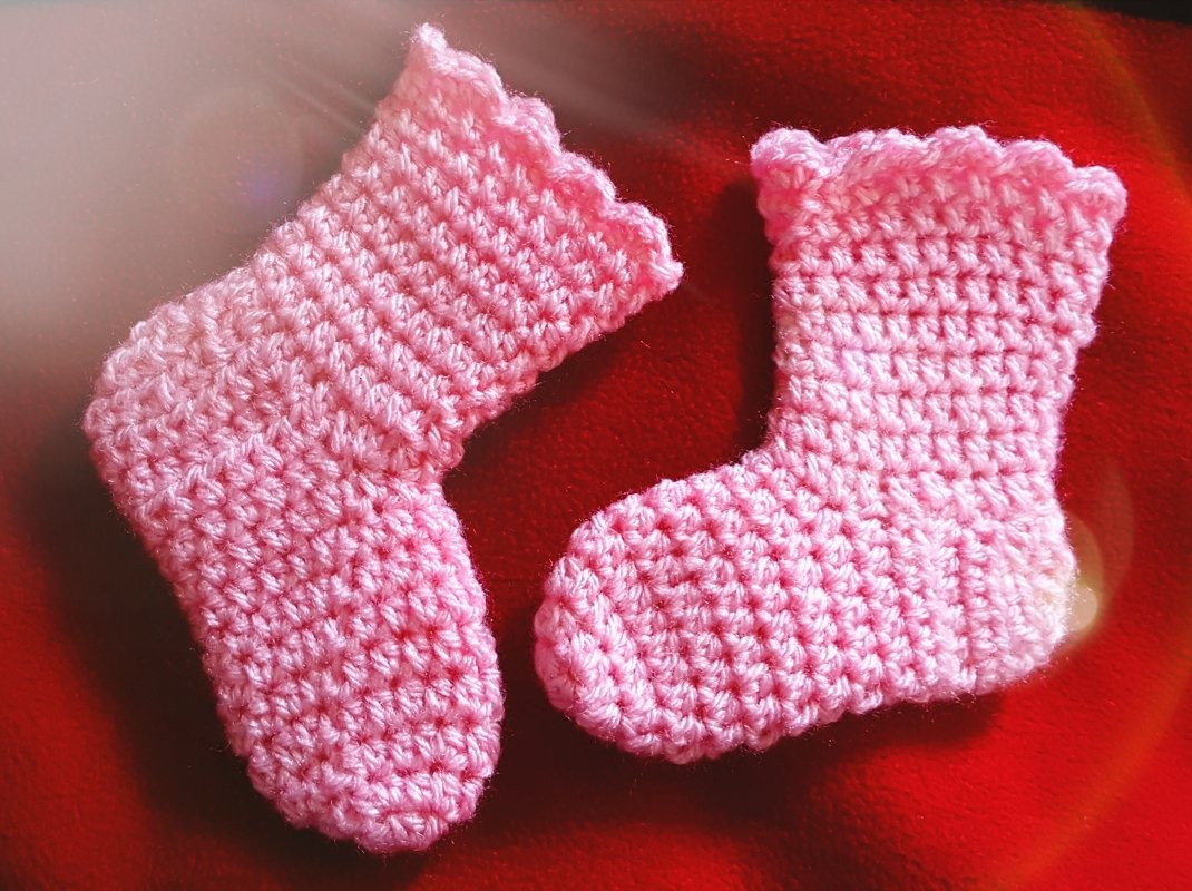 Zwei rosa handgehäkelte Babysocken mit gewelltem Rand auf rotem Stoff.