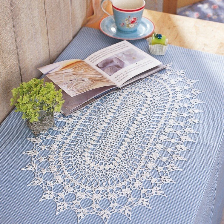 Ovales weißes gehäkeltes Deckchen mit Spitzenmuster auf blau-kariertem Tischläufer, daneben Tasse, Untertasse, offenes Buch und kleine Pflanze