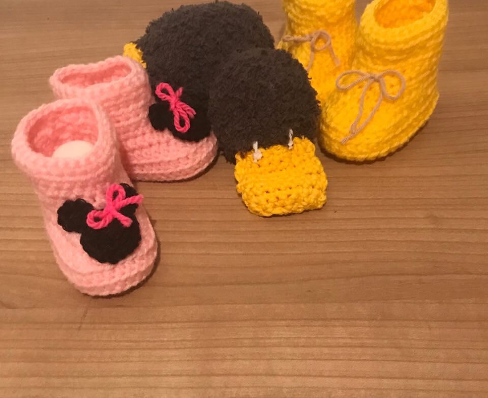 Zwei gehäkelte Babyschuhe: rosa mit schwarzer Applikation und gelb mit Schnürband auf Holzuntergrund