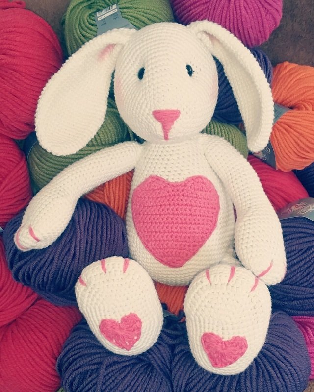 Weißes gehäkeltes Amigurumi-Häschen mit rosa Herz auf der Brust, rosa Nase und Herzchen auf den Pfoten, sitzend auf bunten Wollknäueln.