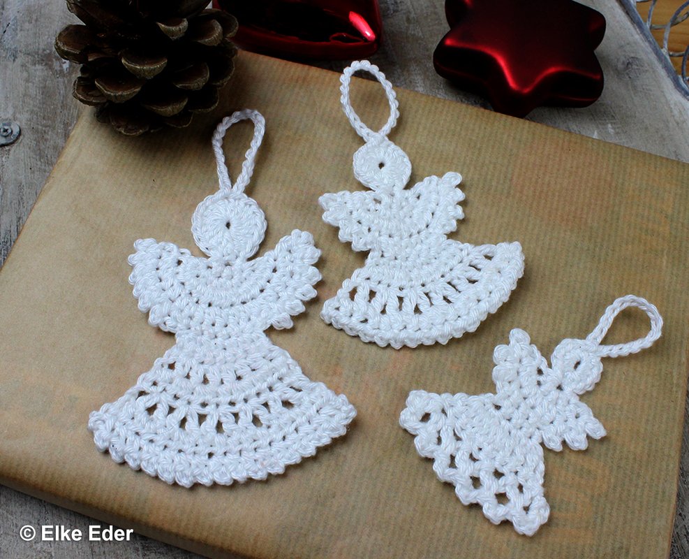 Drei weiße gehäkelte Engel mit Schlaufen auf braunem Packpapier, dekoriert mit Tannenzapfen und roten Weihnachtssternen.