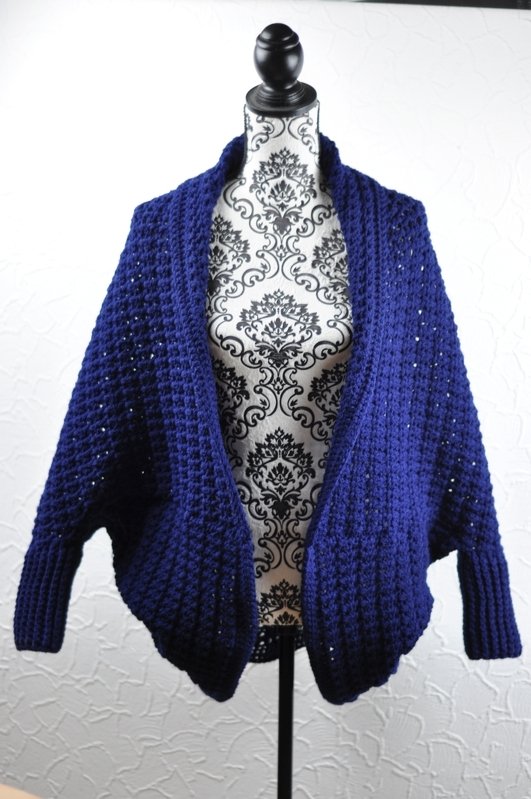 Dark blue crocheted open-front shrug with textured stitch and ribbed cuffs on a mannequin.