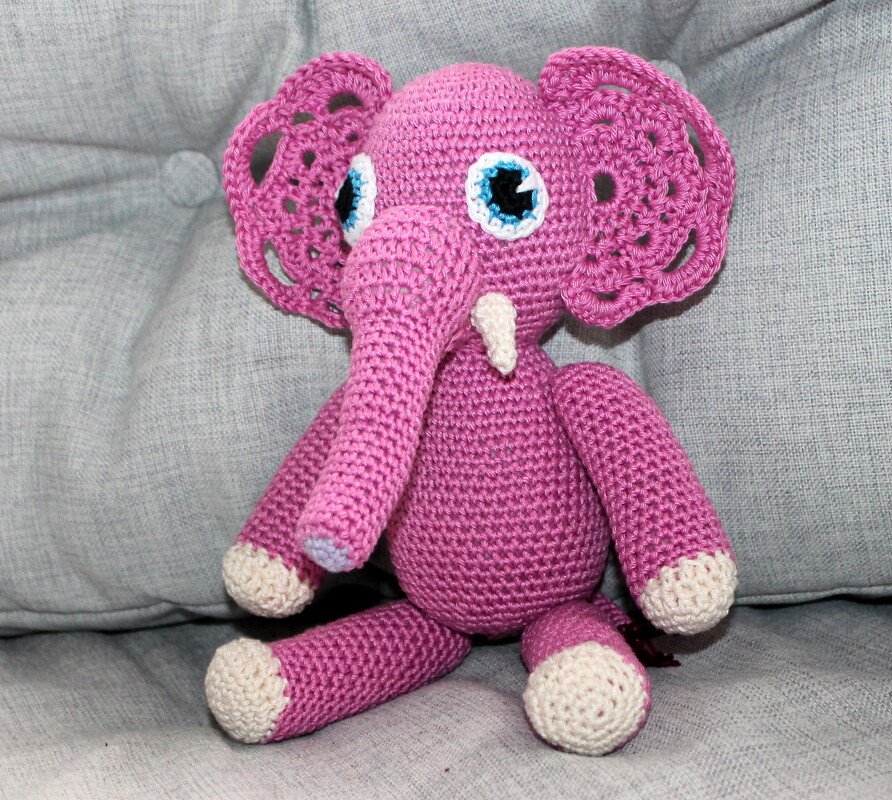 Gehäkelter rosa Amigurumi-Elefant mit hellen Pfoten und applizierten blauen Augen, sitzend auf grauem Sofa