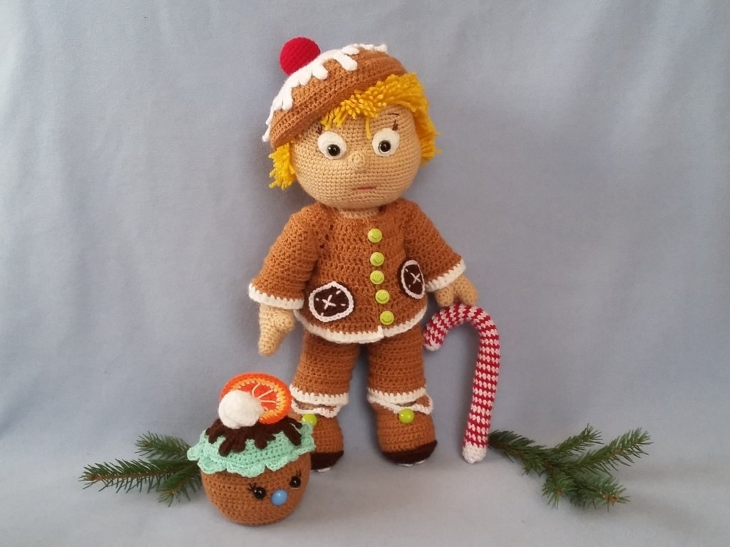 Gehäkelte Amigurumi-Puppe im Lebkuchen-Stil mit brauner Mütze und Jacke, daneben Zuckerstange und Cupcake