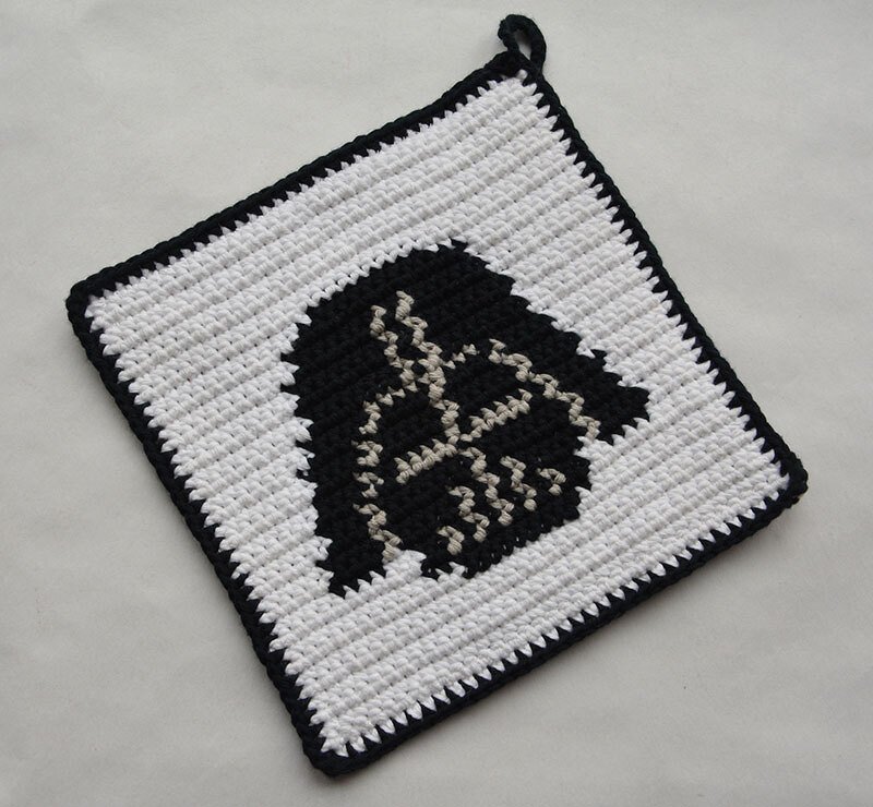 Quadratischer weißer gehäkelter Topflappen mit schwarzer Helm‑Silhouette, schwarzer Einfassung und Aufhängeschlaufe