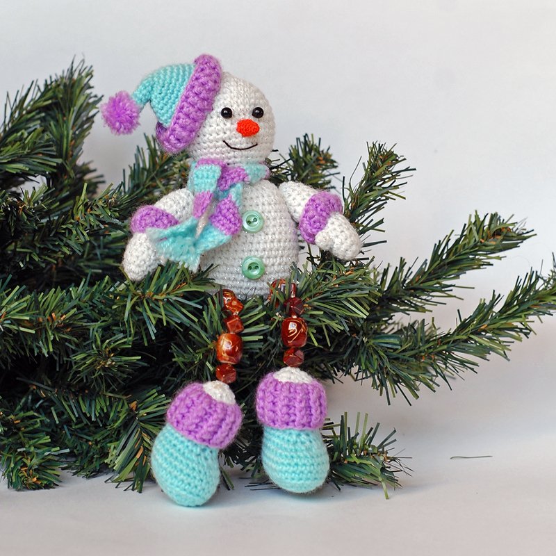Crocheted snowman amigurumi wearing a purple and turquoise striped hat and scarf on pine branches.