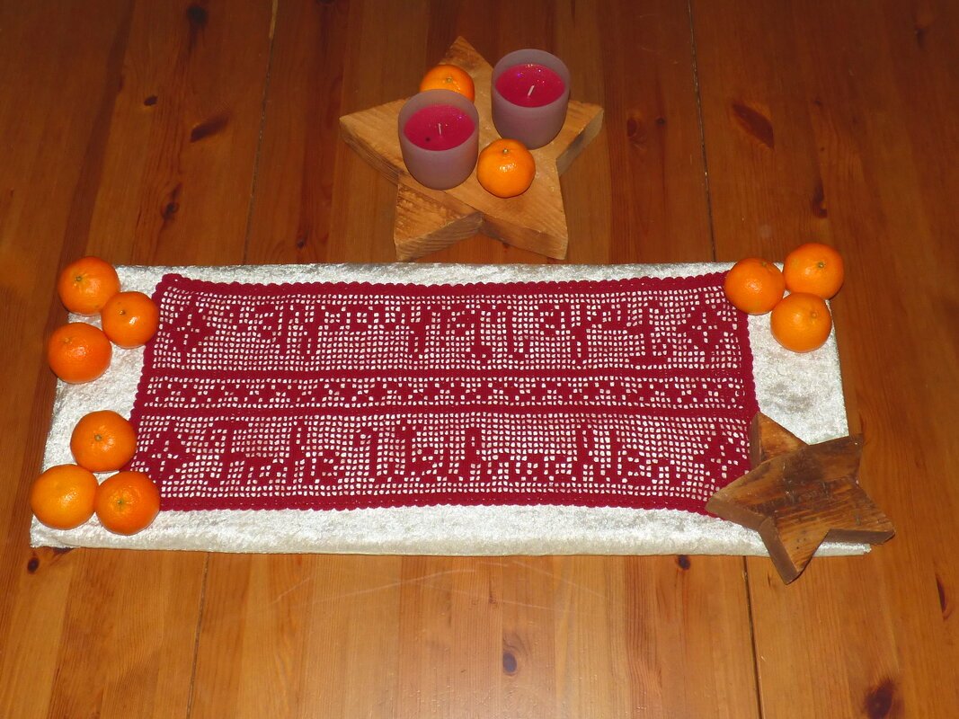 Roter gehäkelter Tischläufer mit den Schriftzügen „Frohe Weihnachten“ und „Schöne Feiertage“, dekoriert mit Mandarinen und Kerzen auf Holzsternen auf einem Holztisch.