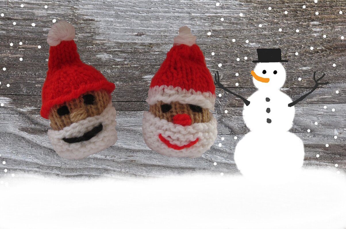 Zwei gestrickte Weihnachtsmannköpfe mit roten Mützen und weißen Bärten vor Holzuntergrund neben gezeichnetem Schneemann