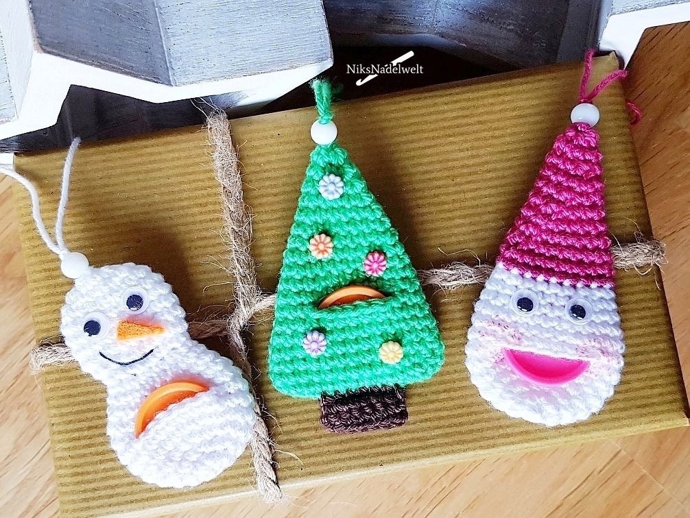 Drei gehäkelte Weihnachts‑Schlüsselanhänger: Schneemann, grüner Tannenbaum und rosafarbener Wichtel auf Geschenkverpackung