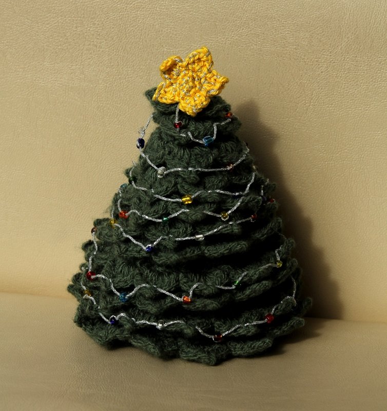 Dunkelgrüner handgehäkelter Mini-Weihnachtsbaum mit gelbem Stern und silberner Perlenkette vor beigem Hintergrund.