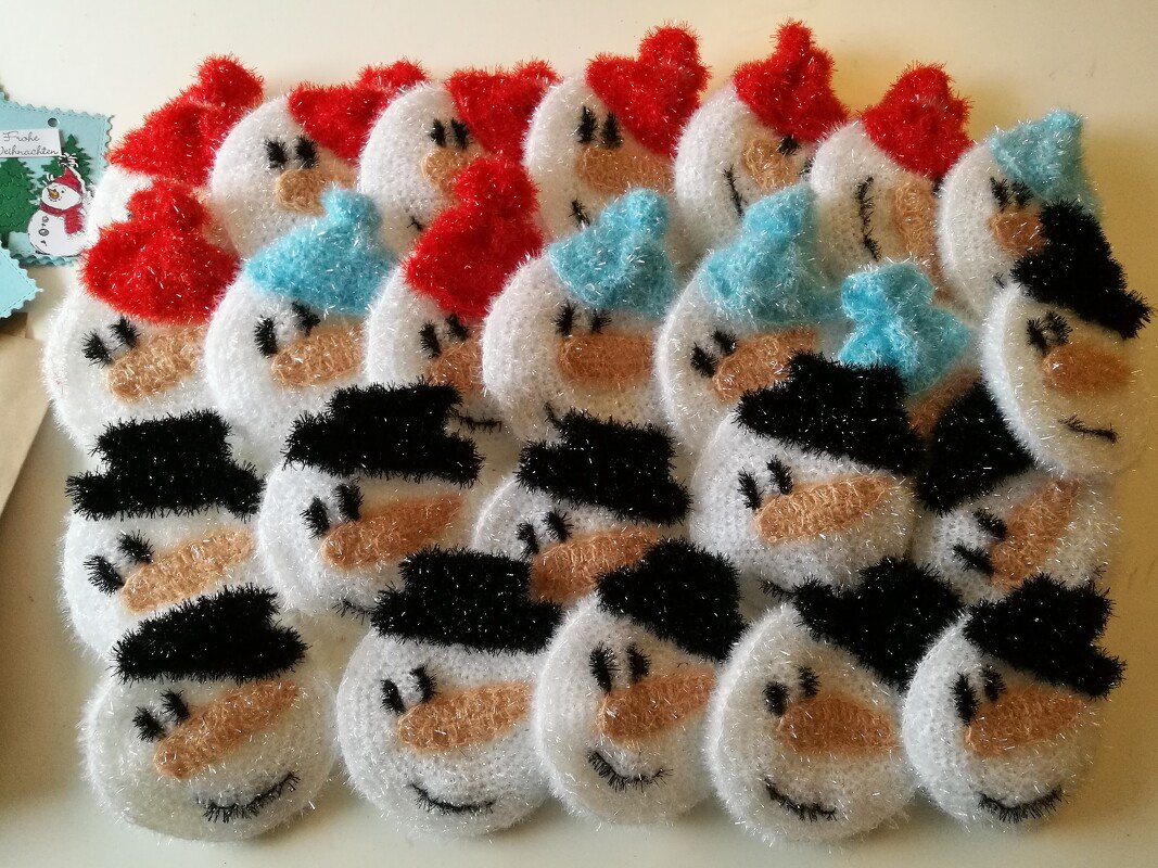 Nahaufnahme von mehreren gehäkelten, runden Schneemann-Gesichtern aus schimmerndem Garn mit verschiedenen Mützenfarben