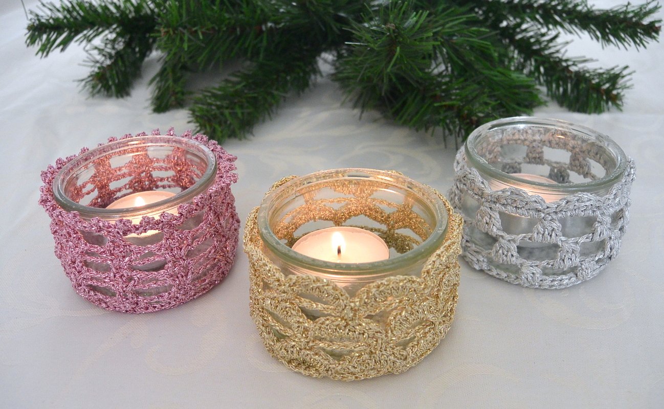 Drei gehäkelte Teelichthalter in Rosa, Gold und Silber mit brennenden Teelichtern und Tannenzweig