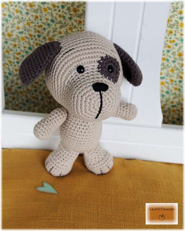 Beiger gehäkelter Amigurumi-Hund mit dunklen Ohren und Augenpatch auf senffarbenem Stoff