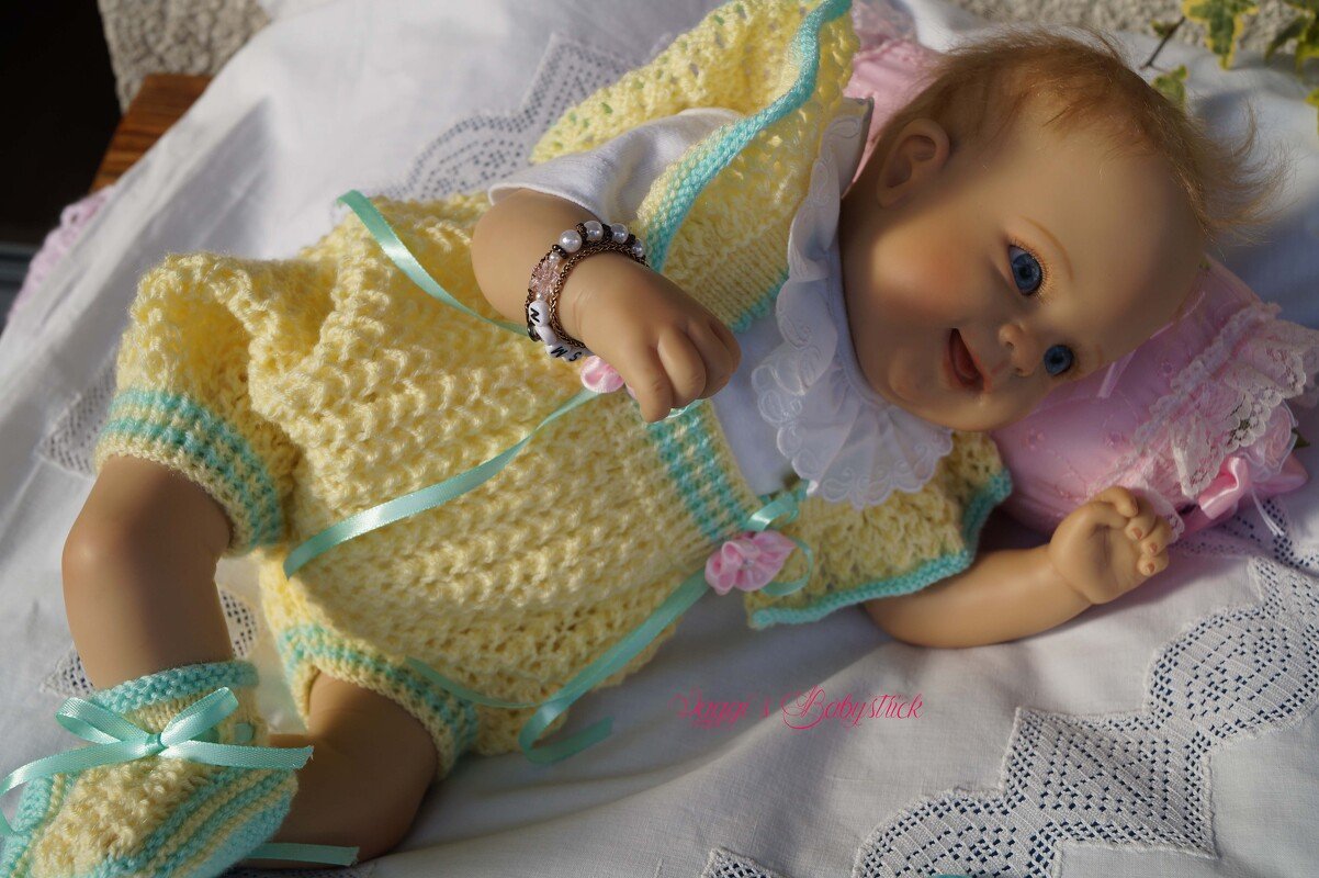 Babypuppe auf weißer Decke trägt ein gelb-hellblau gestricktes Set mit Rüschenkragen, Schühchen und hellgrünen Bändern.