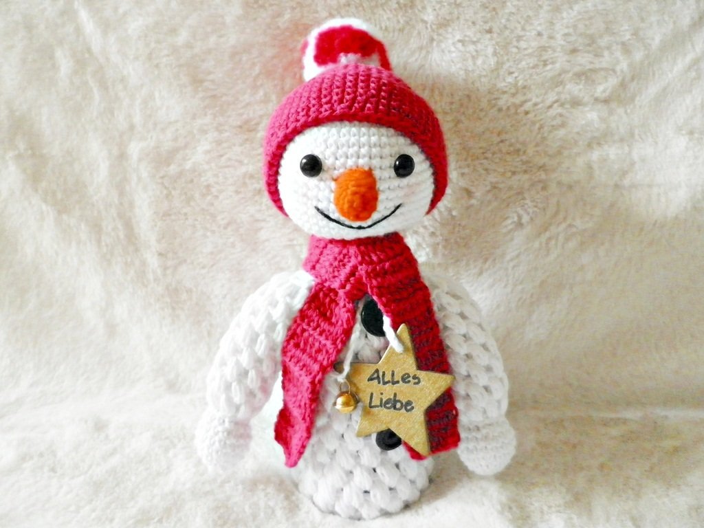 Gehäkelter weißer Schneemann mit roter Mütze und Schal, Holzstern 'Alles Liebe' und Glöckchen auf flauschigem Untergrund.