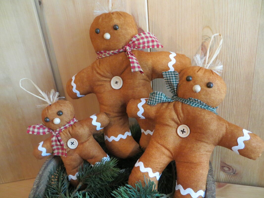 Drei genähte Lebkuchen-Männchen aus braunem Stoff mit weißen Zierborten, karierten Schals, Holzknöpfen, Holzperlen als Nasen und Raffia-Schleifen, präsentiert in einer Schale mit Tannenzweigen vor Holzpaneelen.