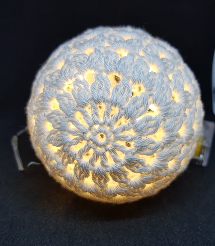 Nahaufnahme einer weiß gehäkelten, von innen leuchtenden Kugel mit filigranem Blumenmuster vor dunklem Hintergrund.