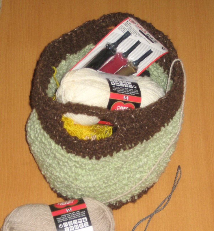 Light green and brown crochet basket holding white yarn and a pack of three crochet hooks, with a beige skein nearby.