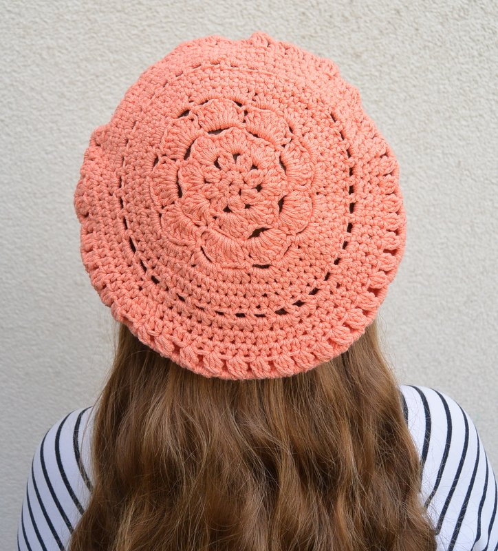 Rückansicht: korallfarbene, handgehäkelte Baskenmütze mit zentralem Blumenmotiv; langes braunes Haar sichtbar