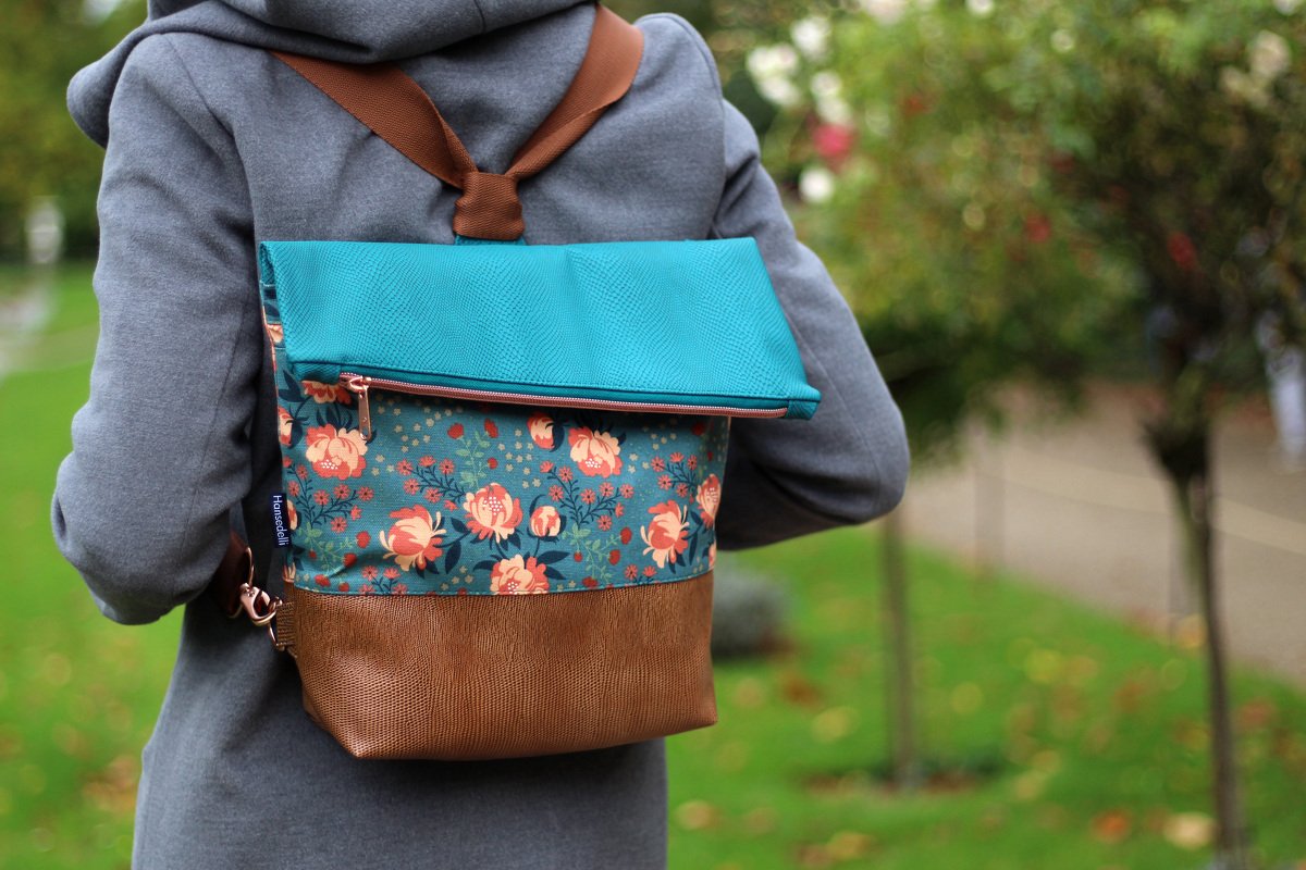 Kleiner Rucksack mit türkiser Klappe, Blumenstoff und braunem, strukturiertem Boden, getragen von einer Person im grauen Mantel