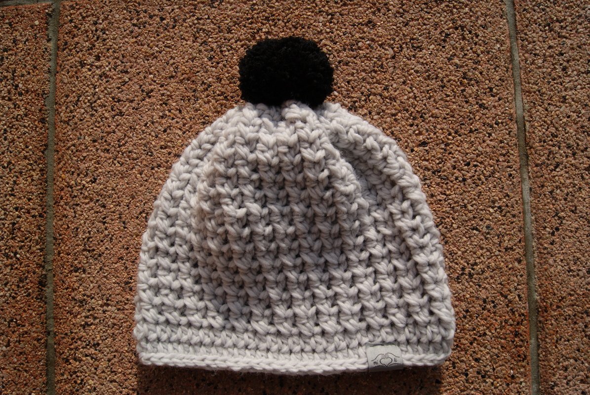Hellgraue gehäkelte Beanie mit schwarzem Bommel auf Steinboden