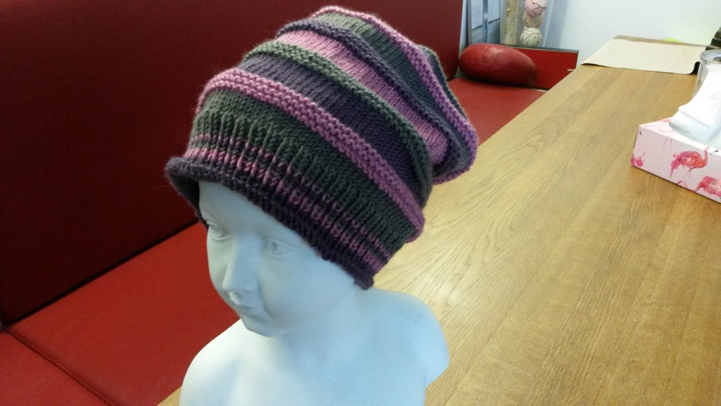 Gestrickte Beanie mit Lila-, Rosa- und Grünstreifen auf weißer Schaufensterpuppe vor rotem Sitzpolster und Holztisch