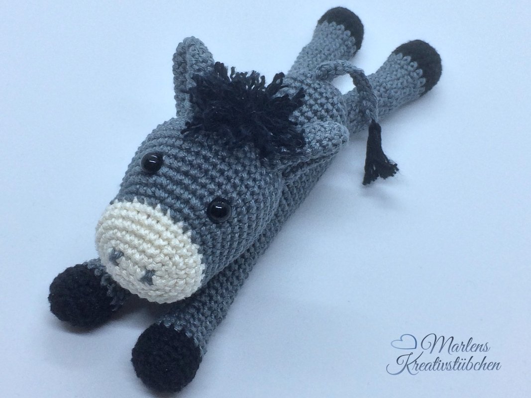 Handgehäkelter grauer Amigurumi-Esel mit hellbeiger Schnauze und schwarzen Hufen, liegend auf hellem Untergrund.