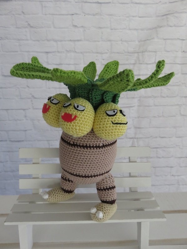 Gehäkelte dreiköpfige Pflanzenfigur (Amigurumi) mit gelben Köpfen und grünen Blättern auf weißer Holzbank vor heller Ziegelwand