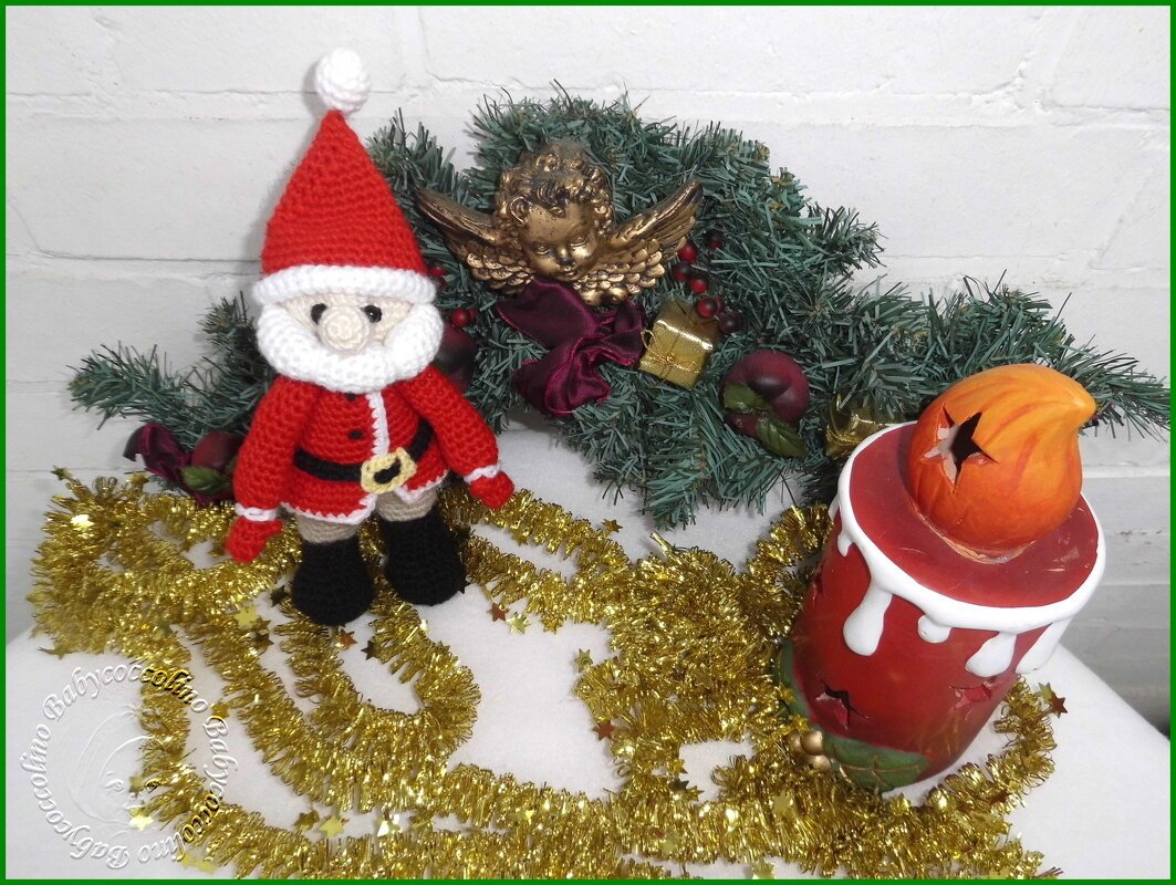 Gehäkelter roter Amigurumi-Weihnachtsmann neben Tannengirlande, goldener Engel-Dekoration und rotem Kerzenhalter