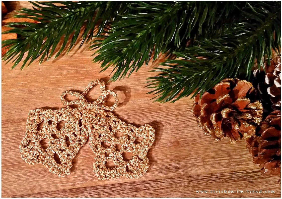 Zwei goldfarbene, gehäkelte Glocken auf einem Holztisch, umrahmt von Tannenzweigen und Tannenzapfen.