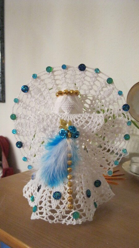 Weißer gehäkelter Weihnachtsengel mit blauen Verzierungen, Federn und goldenen Perlen auf einem Tisch
