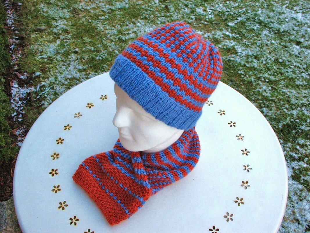 Mannequin mit blau-orange gestreifter gestrickter Mütze und passendem Schlauchschal auf weißem Tisch im Außenbereich