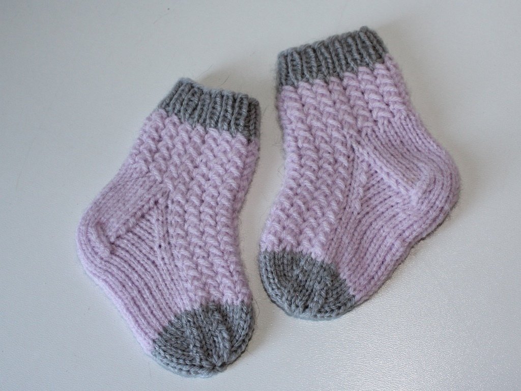 Paar gestrickter Babysocken in Flieder mit grauem Bündchen, Ferse und Spitze auf hellem Untergrund