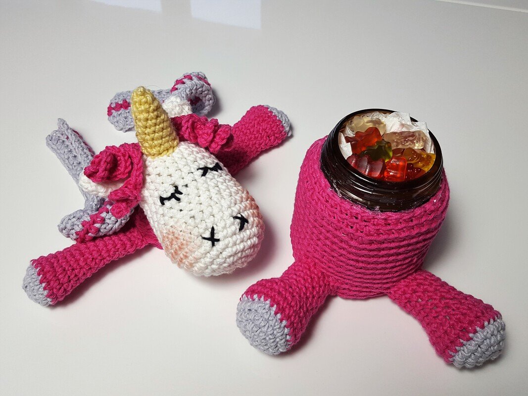 Gehäkeltes Einhorn-Kuscheltier und rosafarbenes Utensilo, das ein Glas mit Gummibärchen hält, vor weißem Hintergrund.