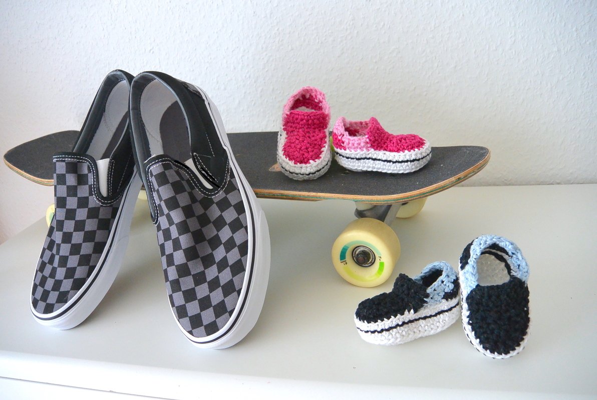 Zwei Paar gehäkelte Babyschuhe (pink, hellblau) neben kariertem Slip
on und Skateboard zum Größen
und Farbvergleich