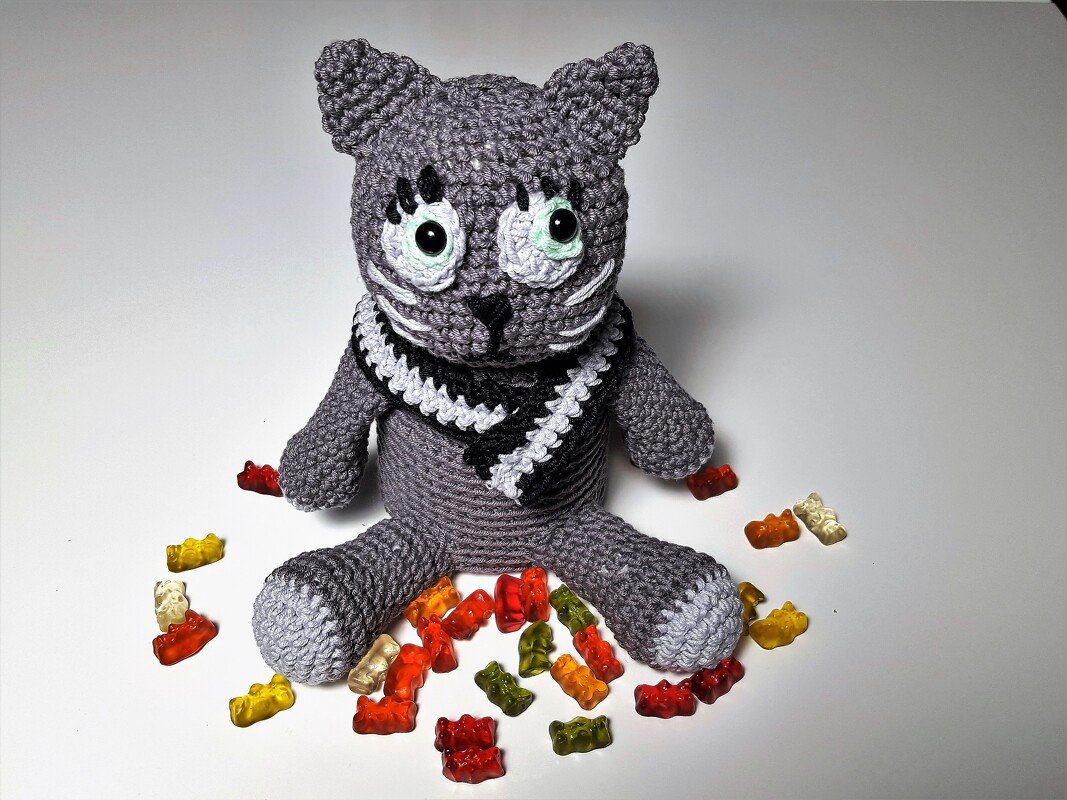 Gehäkelte graue Amigurumi‑Katze mit schwarz-weißem Schal und bunten Gummibärchen