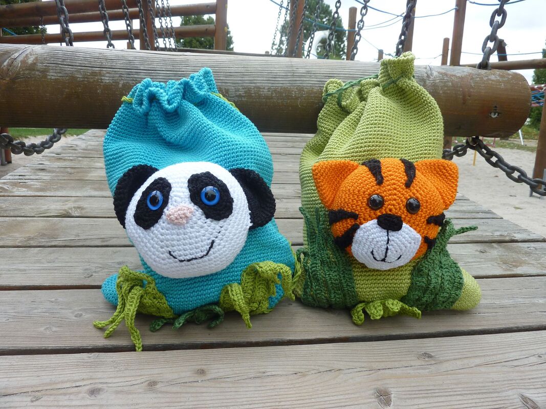 Zwei gehäkelte Kordelbeutel mit applizierten Tiergesichtern (blauer Panda, grüner Tiger) auf Holzplanken