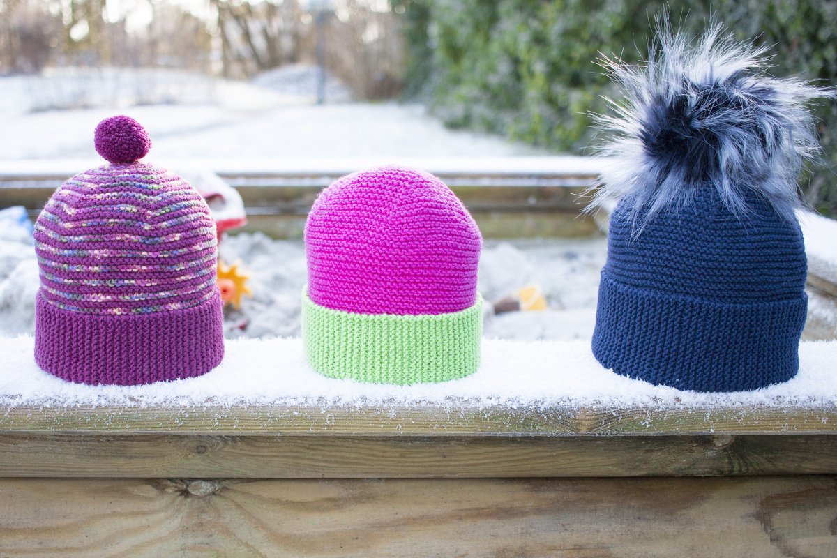 Drei verschiedene gestrickte Mützen auf einem schneebedeckten Holzbrett.
