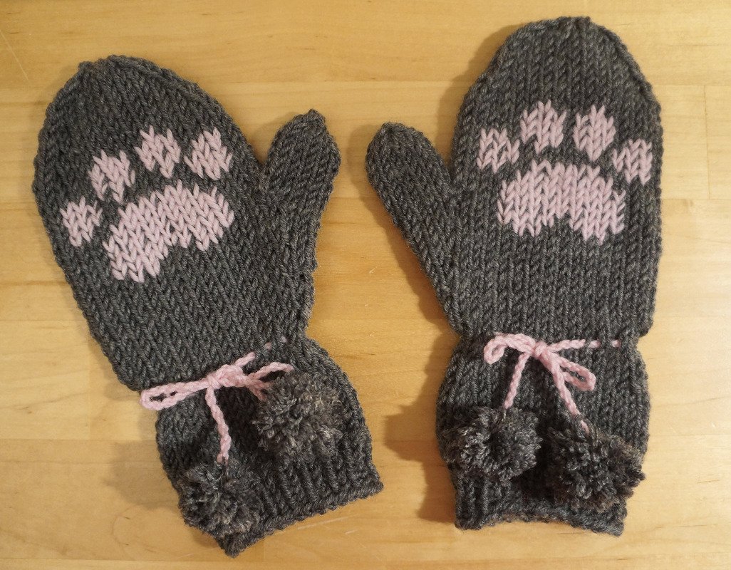 Zwei dunkelgraue Strickfäustlinge mit rosa Pfotenmotiv, rosa Bindebändern und grauen Bommeln auf hellem Holzuntergrund.