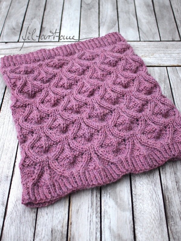 Nahaufnahme eines mauvefarbenen, gestrickten Loop‑Schals mit reliefartigem Muster auf hellem Holzhintergrund.